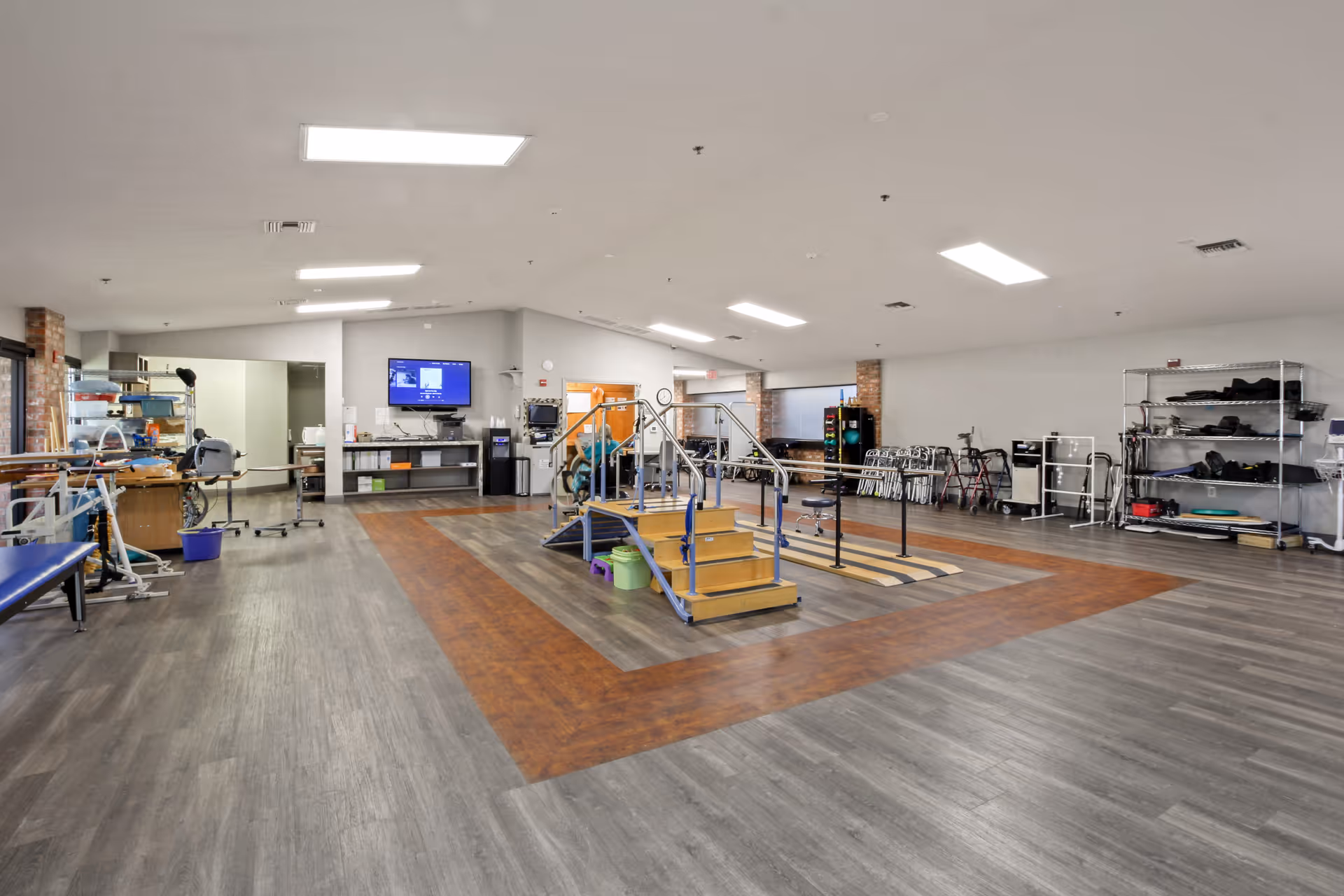 A spacious rehabilitation room with wooden flooring and a central exercise area featuring stairs and handrails for physical therapy. The room contains various medical and therapy equipment, including walkers, chairs, and shelves with supplies. Bright overhead lights illuminate the clean, organized space.