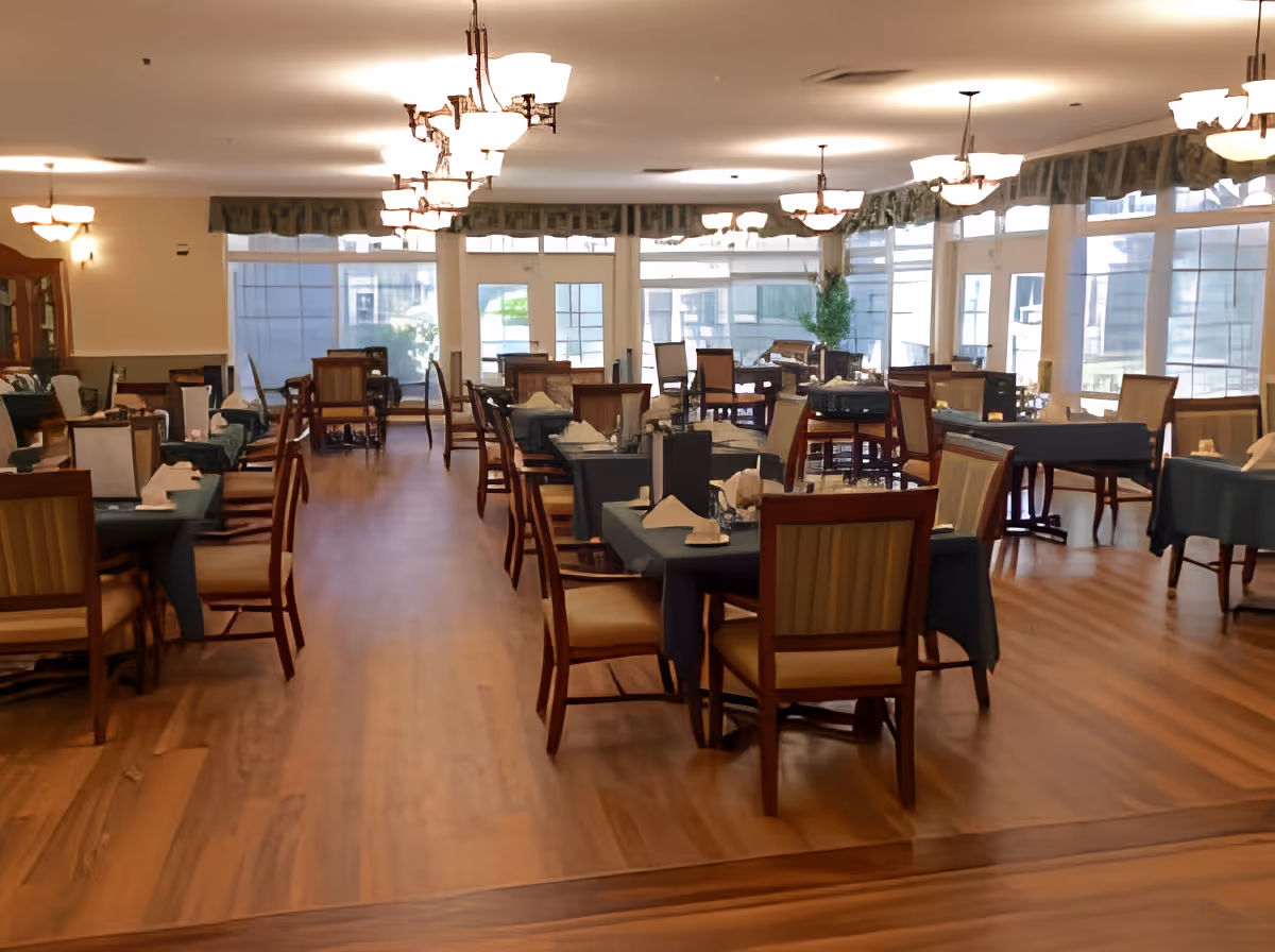 Spacious dining room with multiple tables covered with dark tablecloths, each set with napkins and glassware. Wooden chairs with cushioned seats surround the tables. Large windows line the walls, allowing natural light to fill the room. Ceiling lights hang evenly spaced across the ceiling, and the floor is a light wood finish.