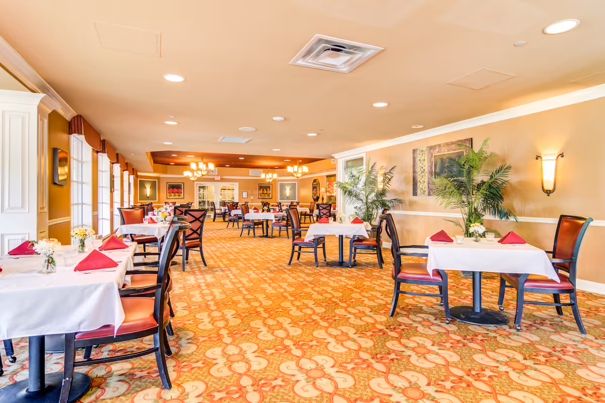 A spacious dining room with multiple tables covered in white tablecloths and set with red napkins and small floral centerpieces. The room features patterned carpet, warm beige walls with framed artwork, large windows with valances, and several chandeliers providing lighting. There are also some potted plants along the walls.