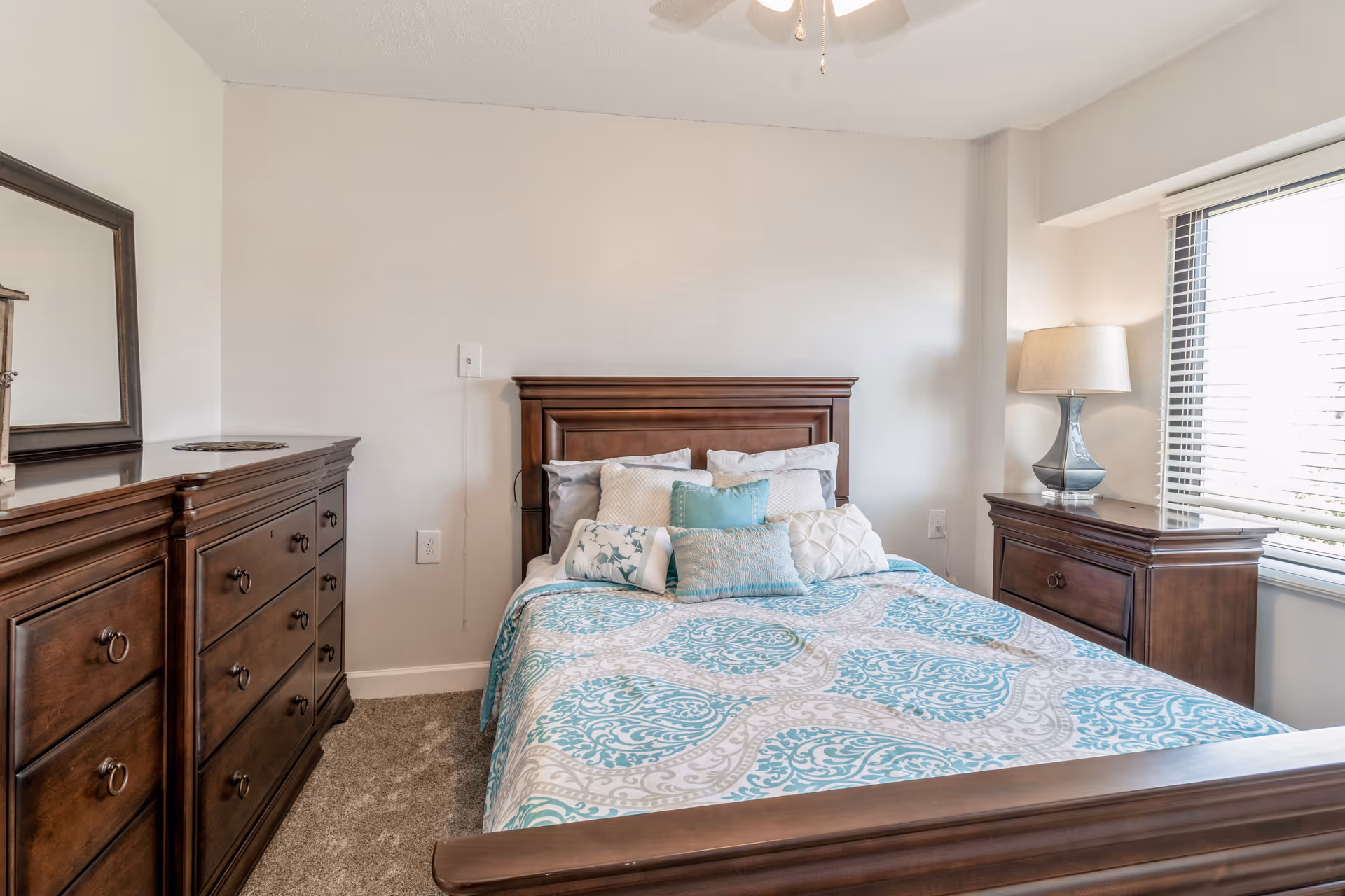 A bedroom with a wooden bed frame and headboard, a bed made with a patterned blue and white bedspread and several decorative pillows. To the left is a wooden dresser with a large mirror on top, and to the right is a wooden nightstand with a lamp. A window with blinds is on the right wall, allowing natural light into the room.