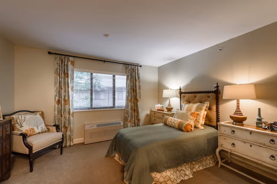 A cozy bedroom in a senior living facility featuring a bed with green bedding and multiple pillows, two bedside tables each with a lamp, a cushioned armchair with a decorative pillow, floral curtains framing a window, and neutral-colored walls and carpet.