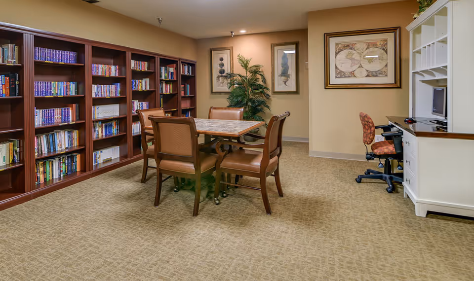 A cozy senior-living common room with bookshelves along the left wall, a central table and chairs, and a computer desk on the right.