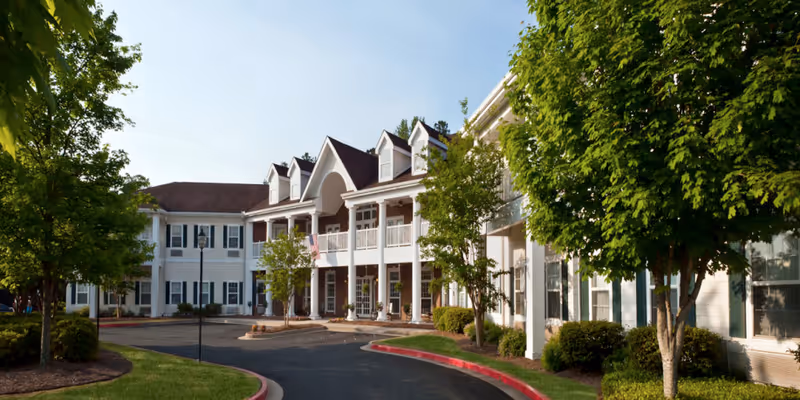 Exterior view of a two-story senior living facility building with white siding, multiple windows, and a covered entrance with columns. The driveway curves in front of the building, and there are trees and shrubs lining the walkway and parking area under a clear blue sky.