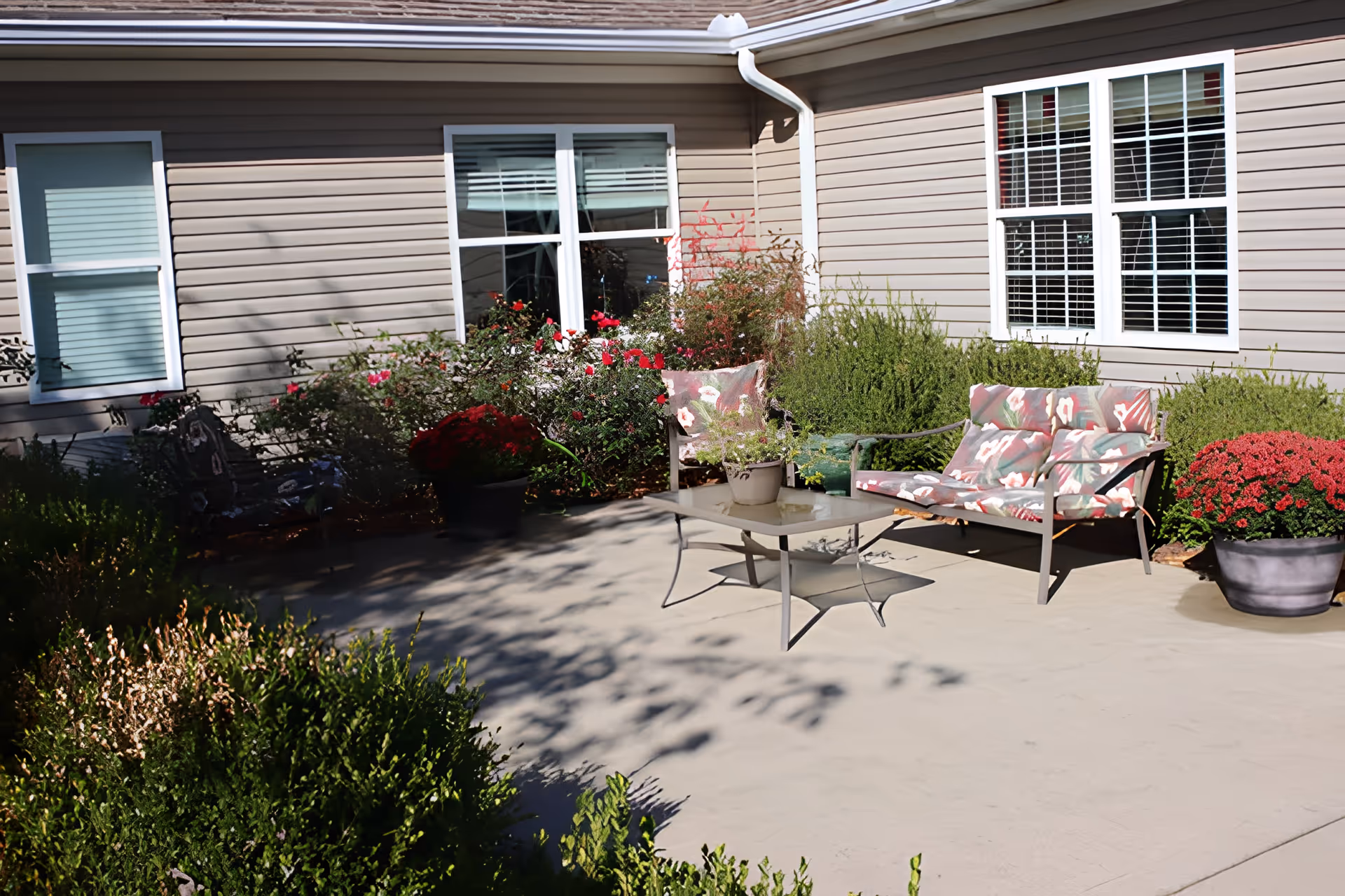Outdoor patio area at Barkley Plantation with cushioned metal chairs and a glass-top table surrounded by various potted plants and bushes, adjacent to a beige siding building with white-framed windows.