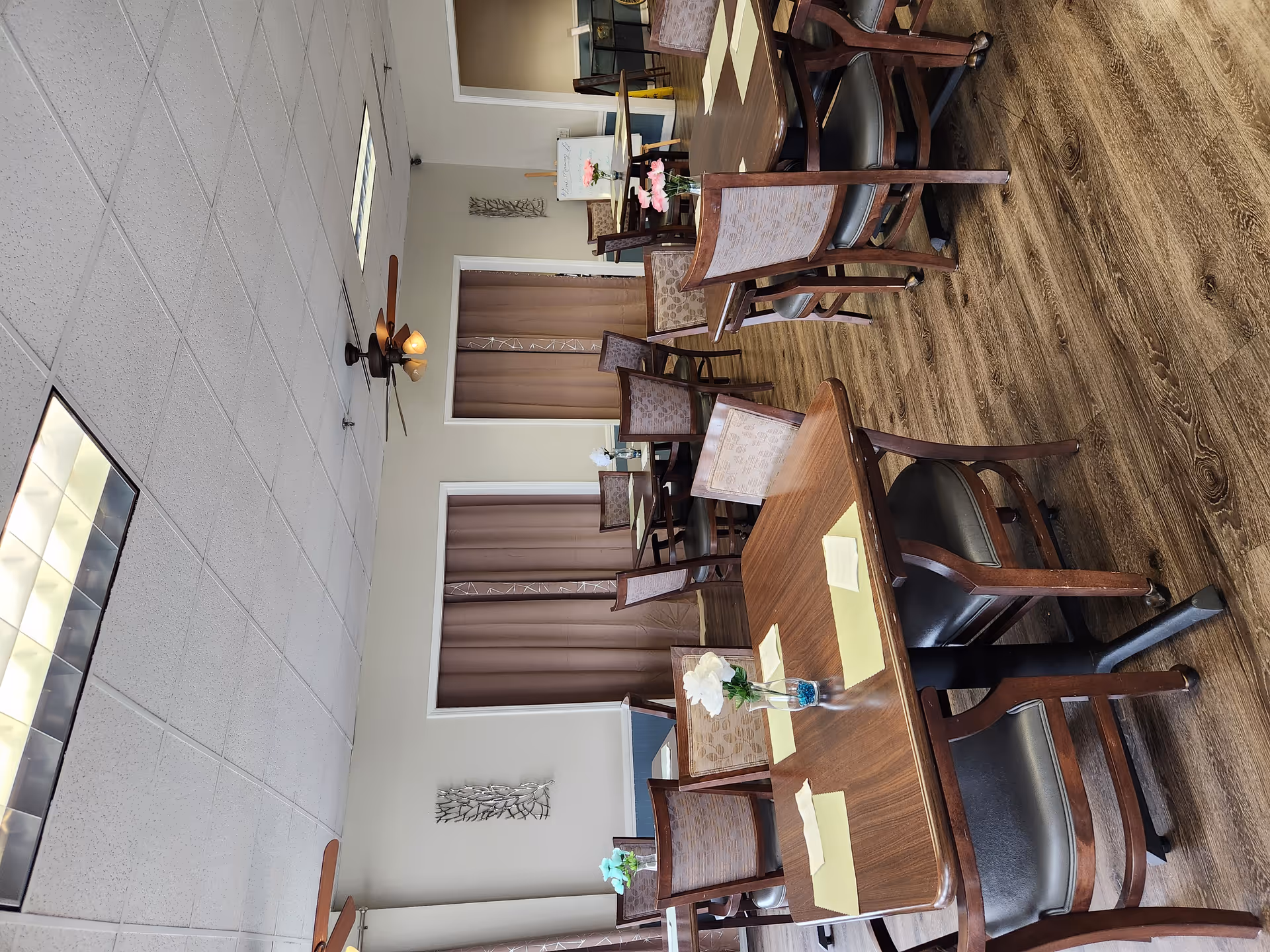 A dining room with several wooden tables and chairs arranged neatly. Each table has placemats and a small vase with flowers. The room has wood flooring, ceiling fans with lights, and beige walls with decorative wall hangings. There are brown curtains covering windows or doorways on one wall.