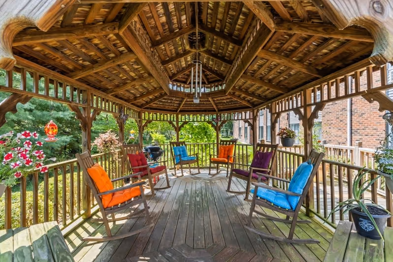 Wooden gazebo with a pitched roof and wooden floor, furnished with rocking chairs that have colorful cushions in orange, red, blue, and purple. Hanging flower pots and decorative bird feeders are attached to the gazebo's railing. The gazebo is surrounded by greenery and is adjacent to a brick building.