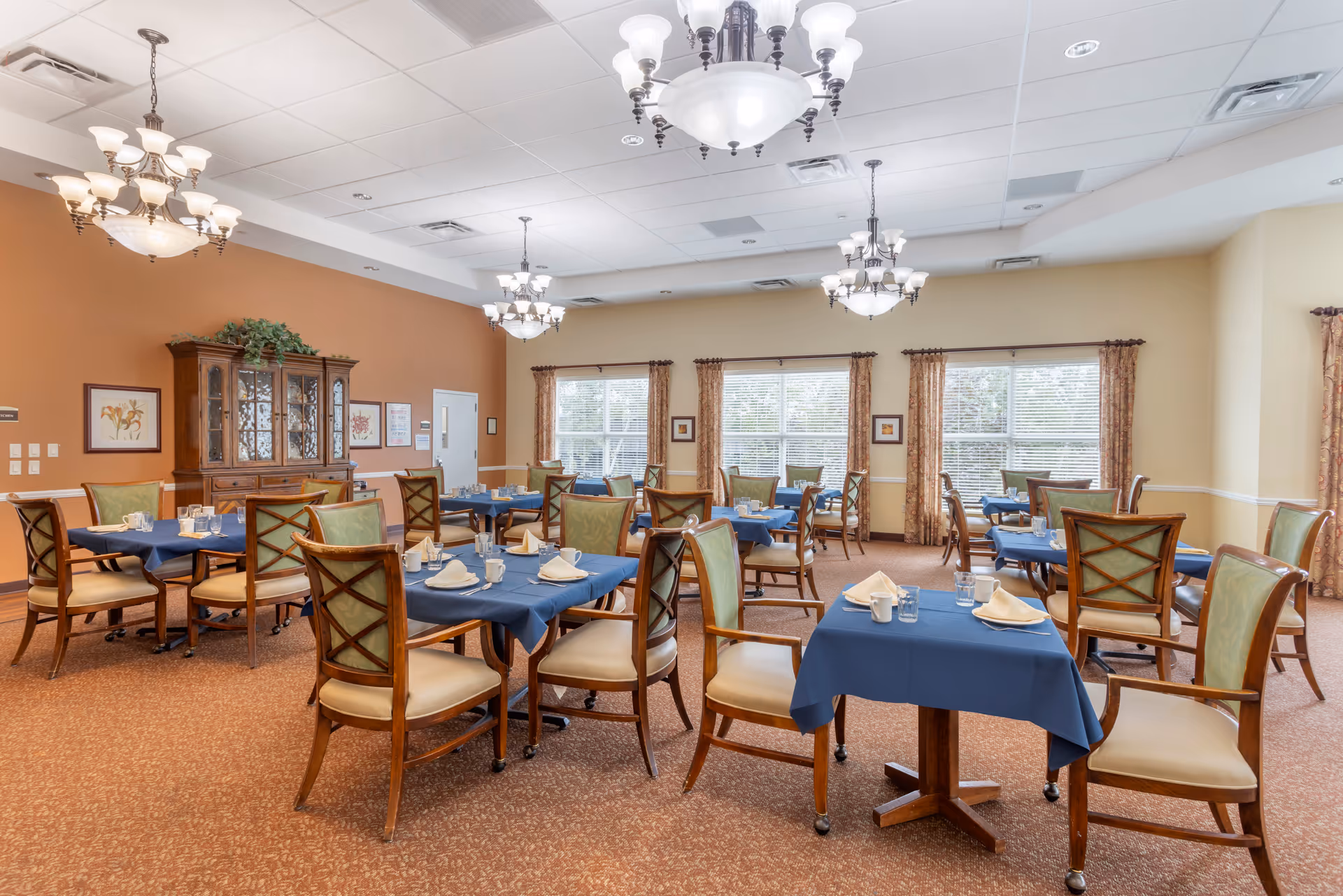 A well-lit dining room with multiple tables covered in blue tablecloths, each set with napkins, glasses, and silverware. The room features wooden chairs with green cushioned backs and beige seats. Large windows with patterned curtains allow natural light to fill the space. Elegant chandeliers hang from the ceiling, and a wooden cabinet with glass doors is positioned against one wall.