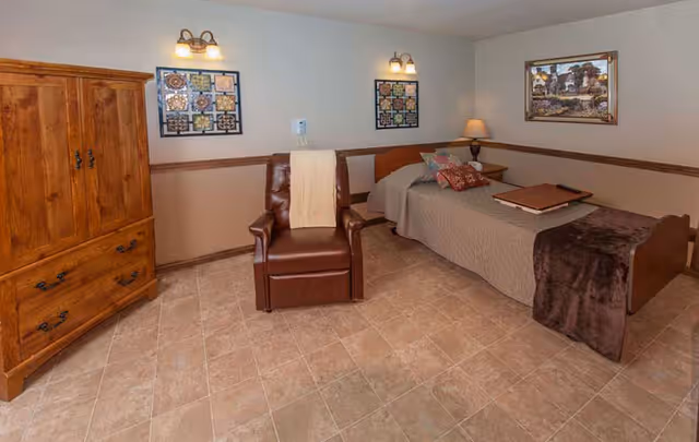A cozy bedroom in a senior living facility featuring a single bed with a brown blanket and pillows, a wooden nightstand with a lamp, a brown leather recliner chair with a light-colored throw draped over it, a large wooden wardrobe, and framed artwork on the beige walls.