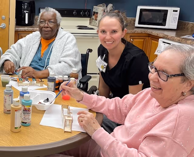 Two elderly women and a caregiver sitting around a table in a kitchen area, engaging in a painting activity with various paint bottles and brushes on the table. The elderly woman on the left is in a wheelchair, and the caregiver is smiling at the camera.