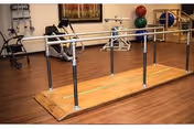 Physical therapy room with parallel bars for walking exercises, exercise balls, and other rehabilitation equipment on a wooden floor.