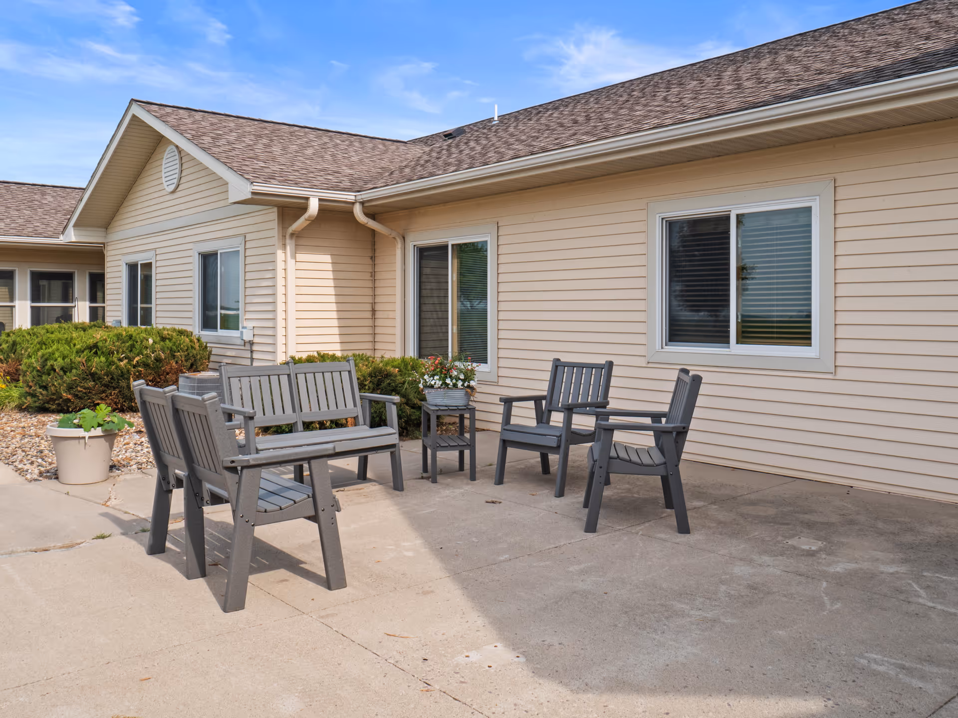 Outdoor patio area at Good Samaritan Society - St. James - Brandt Ridge with gray wooden benches and chairs arranged around a small table with a flower pot, beige siding building with windows in the background, and some bushes and plants along the building.