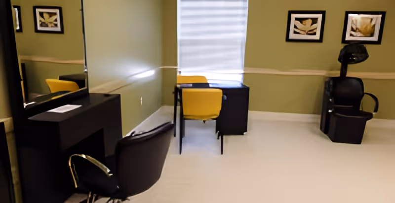 Interior room with light green walls featuring two black salon chairs, a black vanity with a mirror, a black desk with two yellow chairs, and framed artwork on the walls.