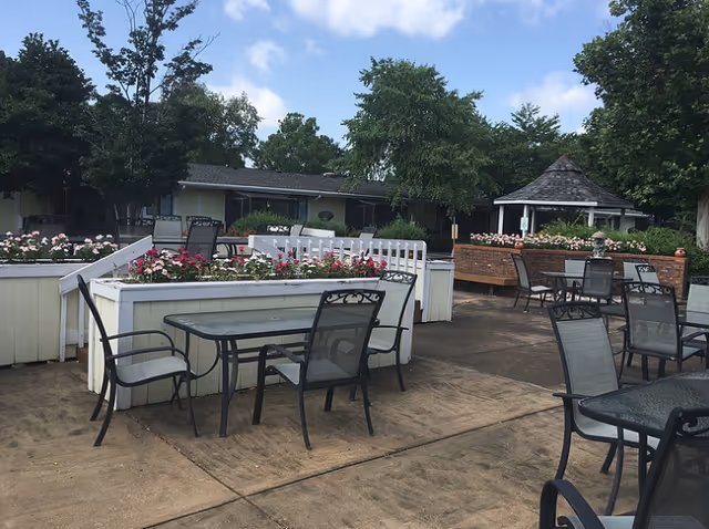 Outdoor patio area with multiple glass-top tables and black metal chairs arranged around raised flower beds filled with pink and red flowers. Trees and shrubs surround the area, and a small gazebo is visible in the background under a partly cloudy sky.