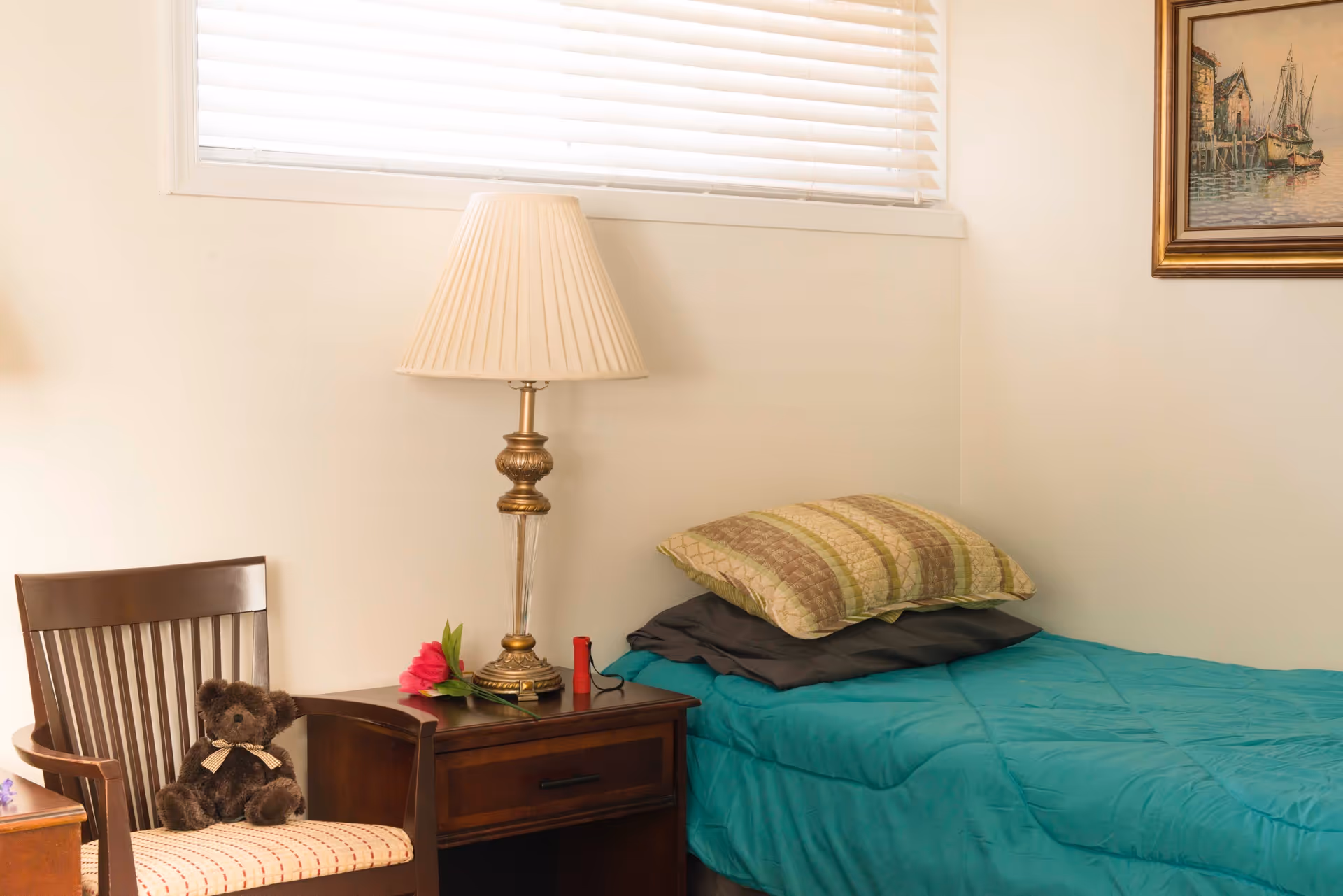 A cozy bedroom corner with a single bed covered in a teal quilt and two pillows, a wooden nightstand with a classic lamp, a red flower, and a small red object. Next to the nightstand is a wooden chair with a teddy bear sitting on it. A window with white blinds and a framed painting of a harbor scene are visible on the wall.
