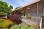 Exterior view of a single-story building with large windows and a covered porch. The porch has a white railing and hanging flower pots. In front of the building, there is a well-maintained garden with various shrubs, bushes, and a tree.