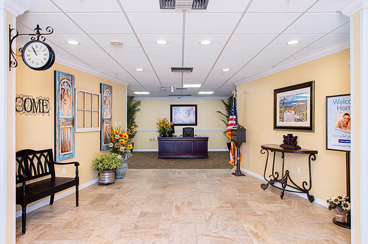 Reception area with a dark wooden desk at the far end, decorated with flower arrangements and an American flag. The walls are painted light yellow and adorned with framed artwork and decorative items. A black bench is placed on the left side under a wall clock and a decorative 'HOME' sign. The floor is tiled with light-colored stone.