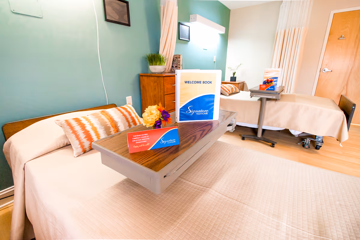 A bright and clean room with two hospital-style beds, each with beige bedding and decorative pillows. Each bed has an overbed table with a welcome book and a small card on it. The walls are painted green and light beige, and there is a wooden dresser with a plant on top. The room has a wooden door and a light-colored floor.