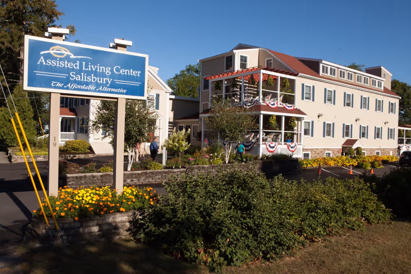 Exterior view of the Assisted Living Center - Salisbury building with a large blue sign in front that reads 'Assisted Living Center Salisbury The Affordable Alternative'. The building is a multi-story light-colored structure with red and white patriotic bunting on the balconies. There are bushes, flowers, and trees in the landscaped area around the building and sign, with a clear blue sky above.