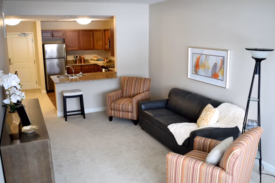 A cozy living room area with a black leather sofa adorned with a white throw blanket and a decorative pillow, two striped armchairs, a tall floor lamp, and a framed abstract painting on the wall. The room has beige carpet and walls, and an open view into a kitchen with wooden cabinets, a stainless steel refrigerator, and a granite countertop with a sink and a stool.