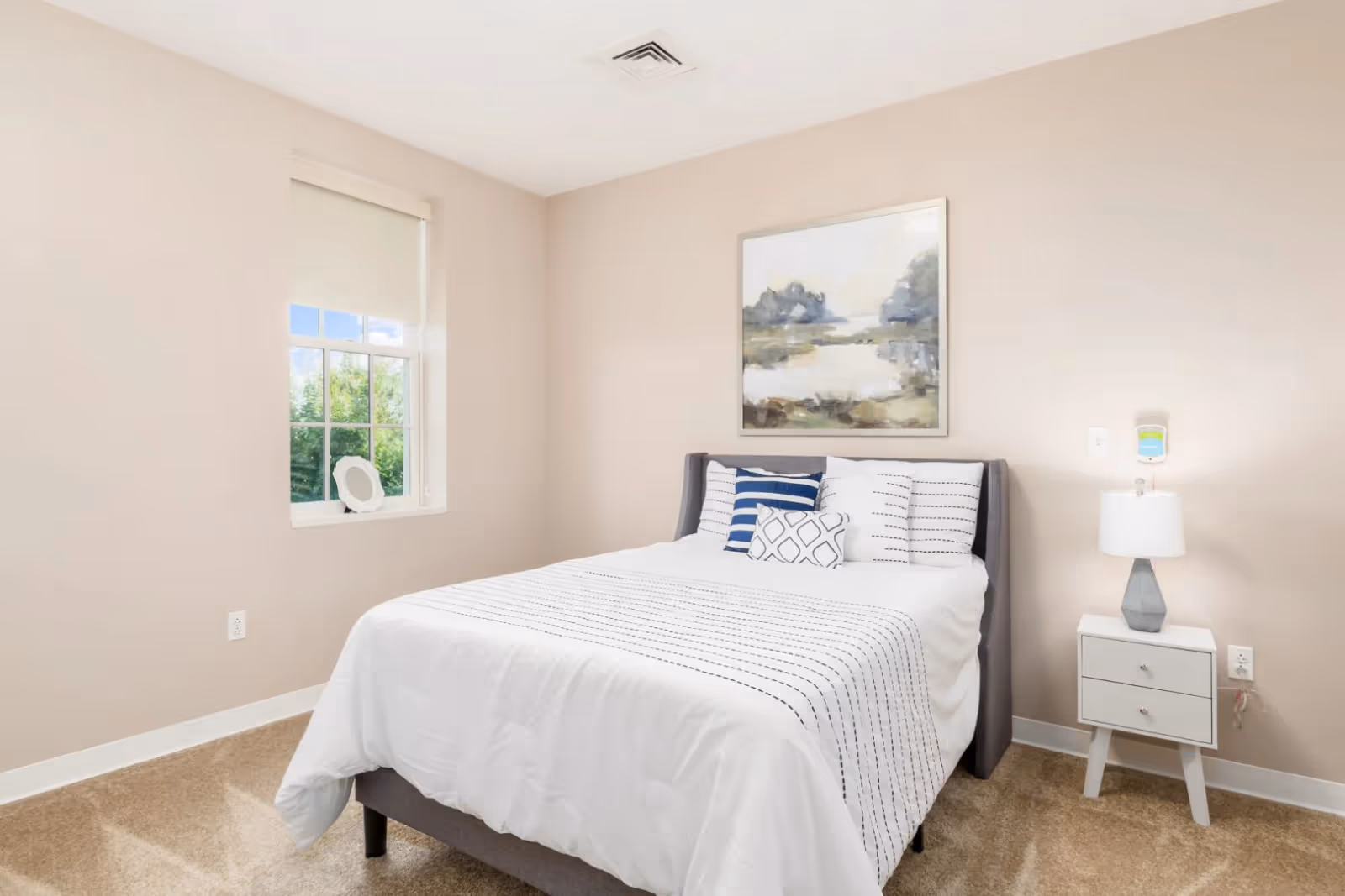 A neatly made bed with white and patterned pillows and a white bedspread with black stripes in a bedroom with beige walls and carpet. There is a window with a white blind partially rolled up, a small white nightstand with two drawers, a modern lamp, and a framed landscape painting hanging above the bed.
