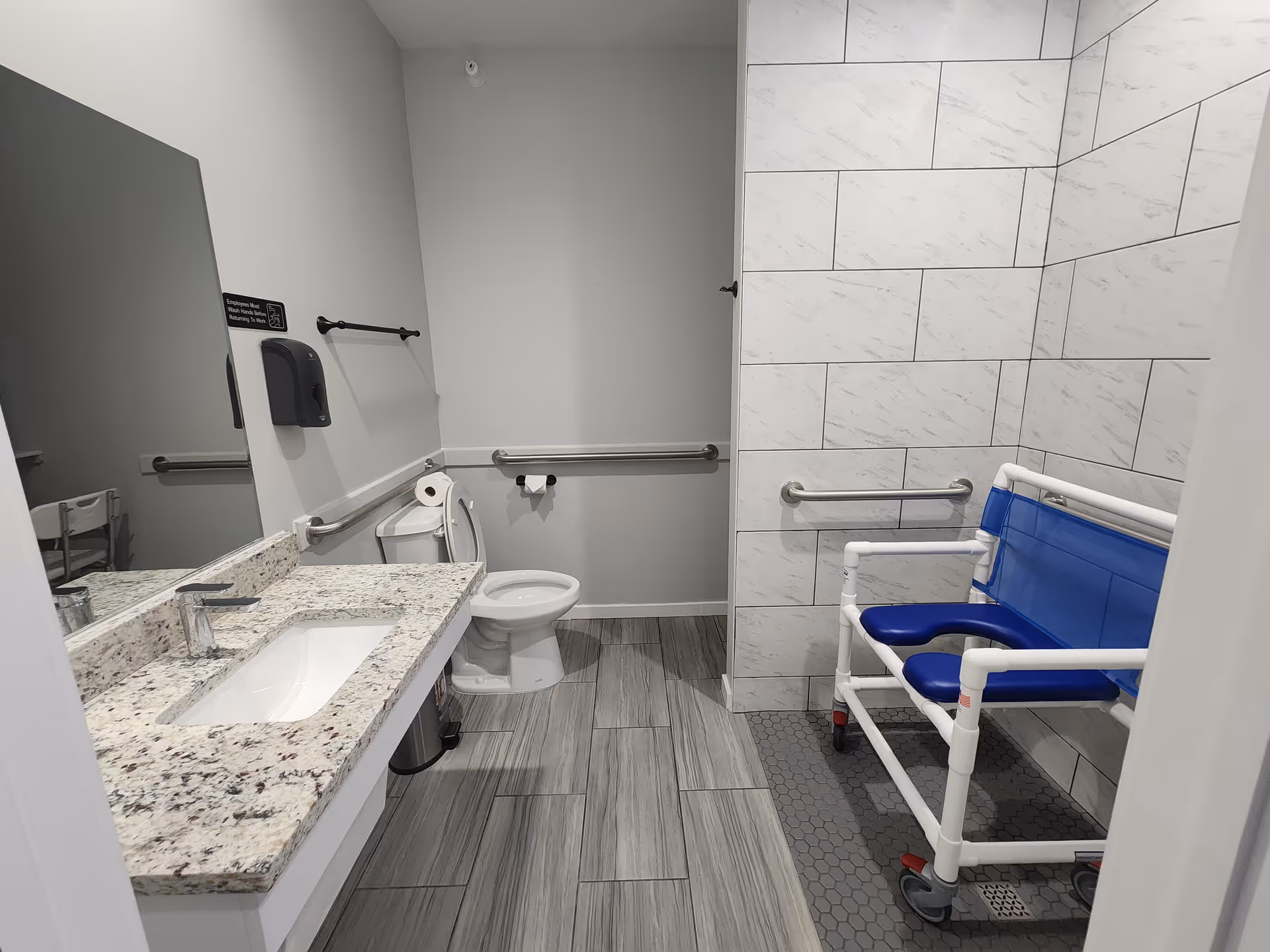 Accessible bathroom with a granite sink countertop, toilet with grab bars, and a blue wheeled shower chair in a tiled shower area.