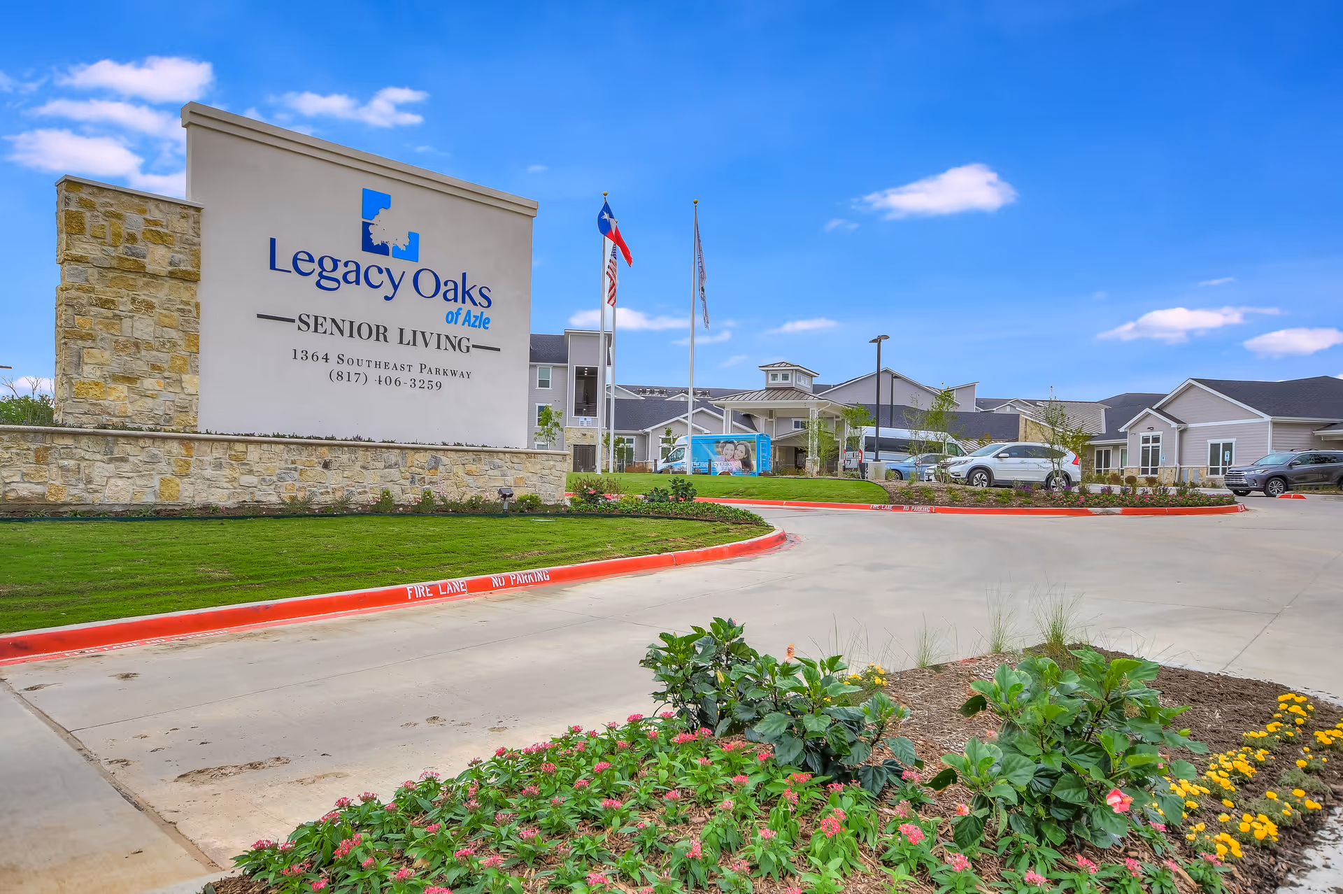 Entrance sign and driveway for Legacy Oaks of Azle senior living with landscaped flower beds and the facility building in the background.
