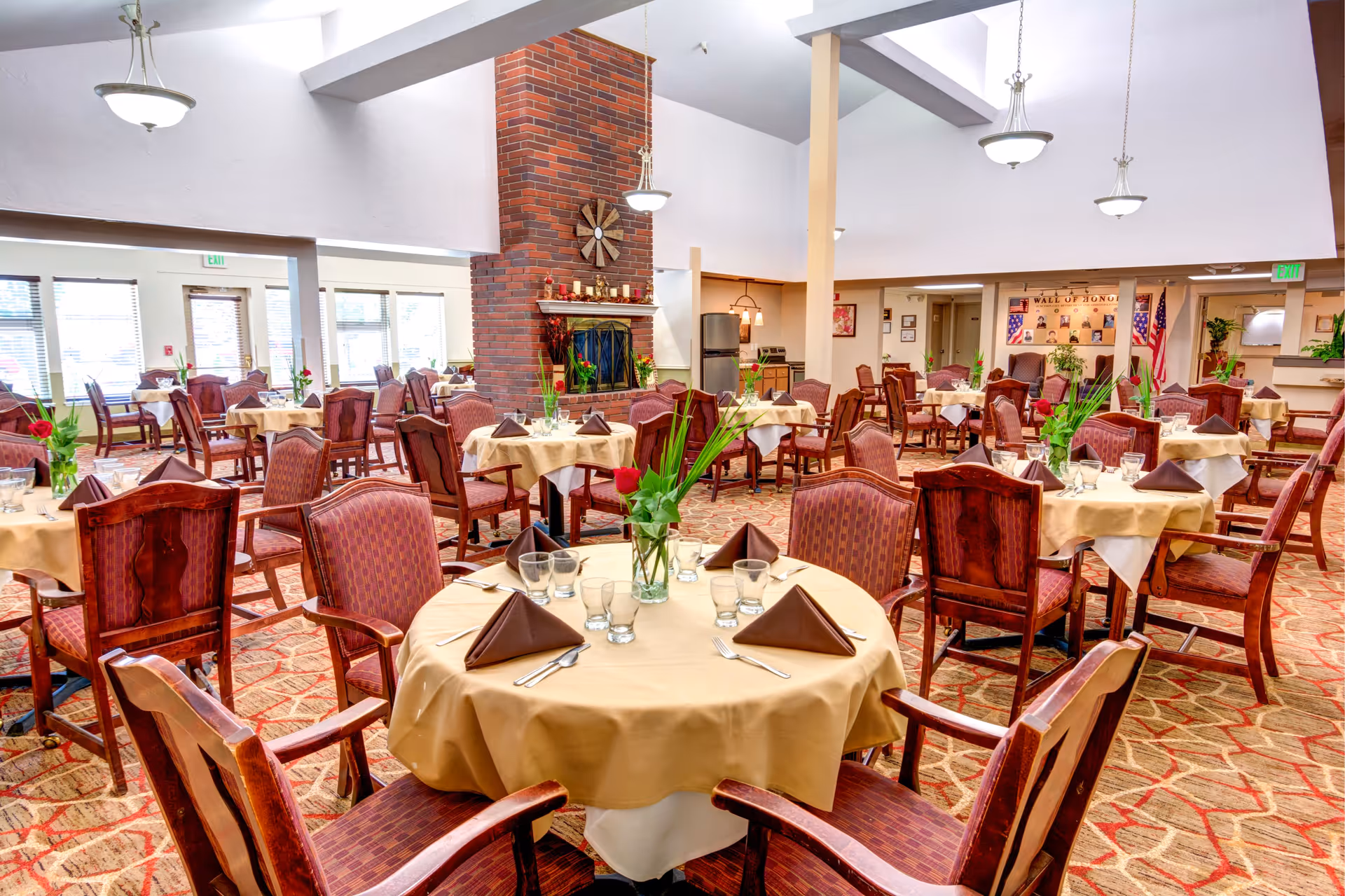Spacious dining room with round tables set with beige tablecloths, folded brown napkins, glassware and wooden chairs around a central brick fireplace.