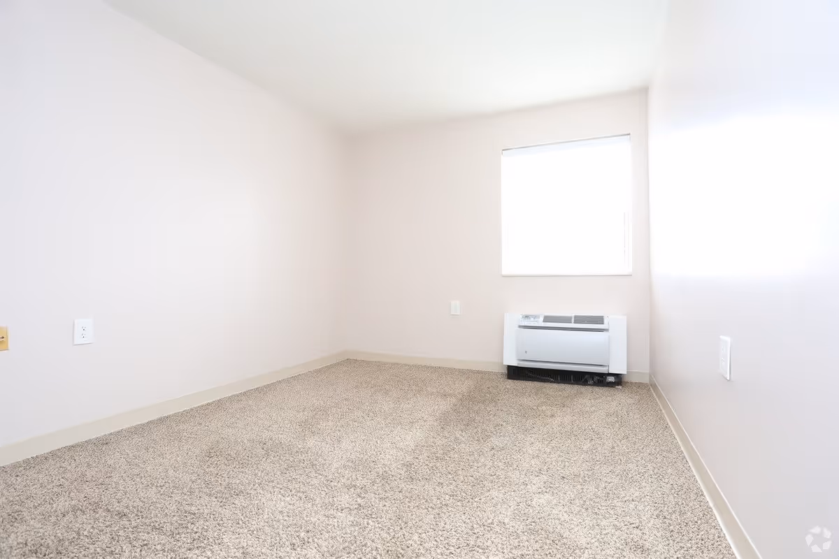Empty small room with beige carpet, light beige walls, a window with blinds, and a wall-mounted air conditioning unit below the window.