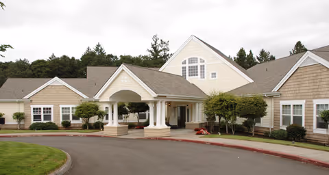 Exterior view of a single-story senior living facility building with beige siding and white trim. The entrance features a covered driveway with white columns and a peaked roof. There are small trees and shrubs planted around the building, and a curved driveway in front.