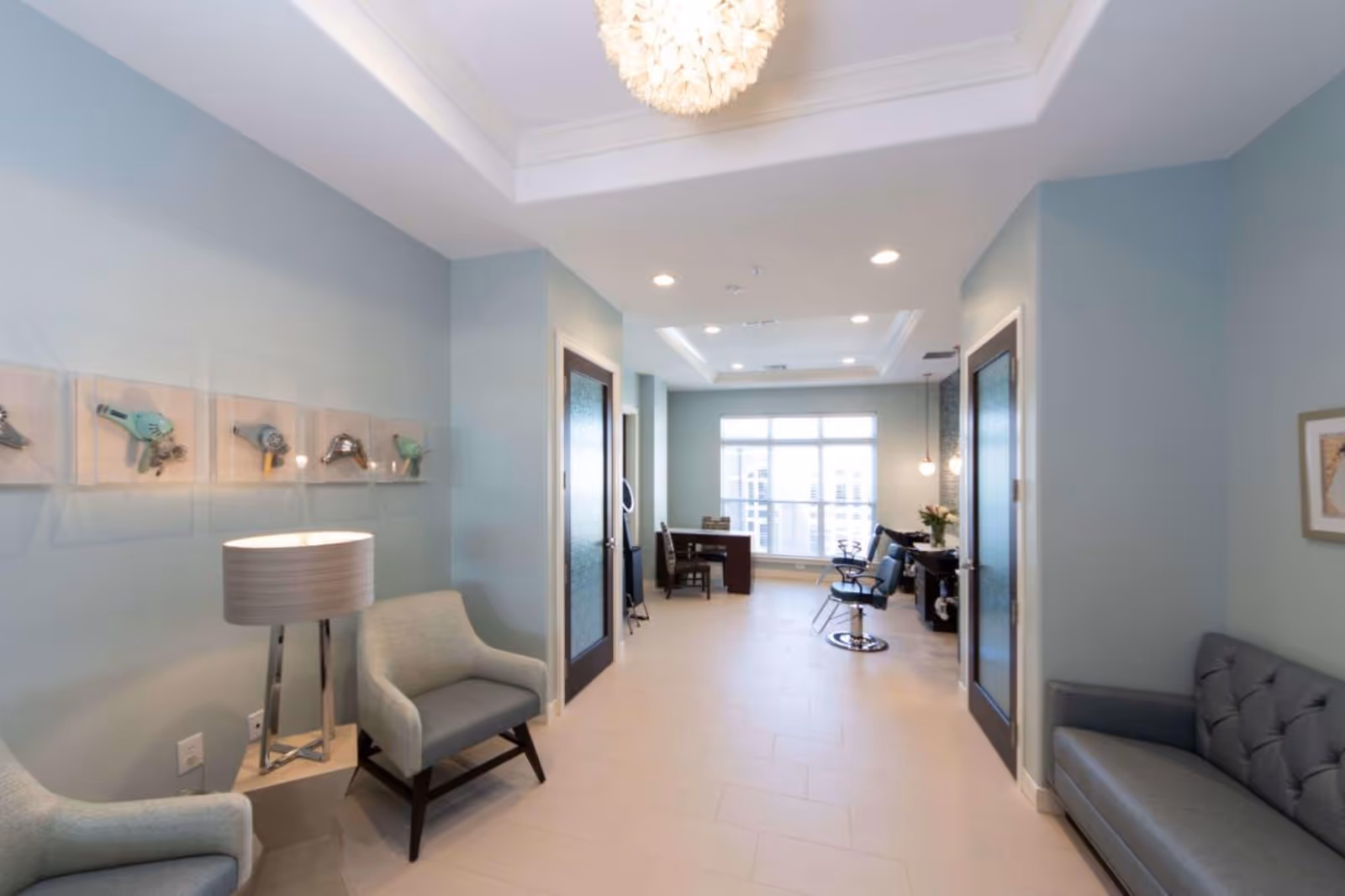 Bright lobby-like seating area in a senior living facility with armchairs, a lamp, and salon stations visible down a hallway.