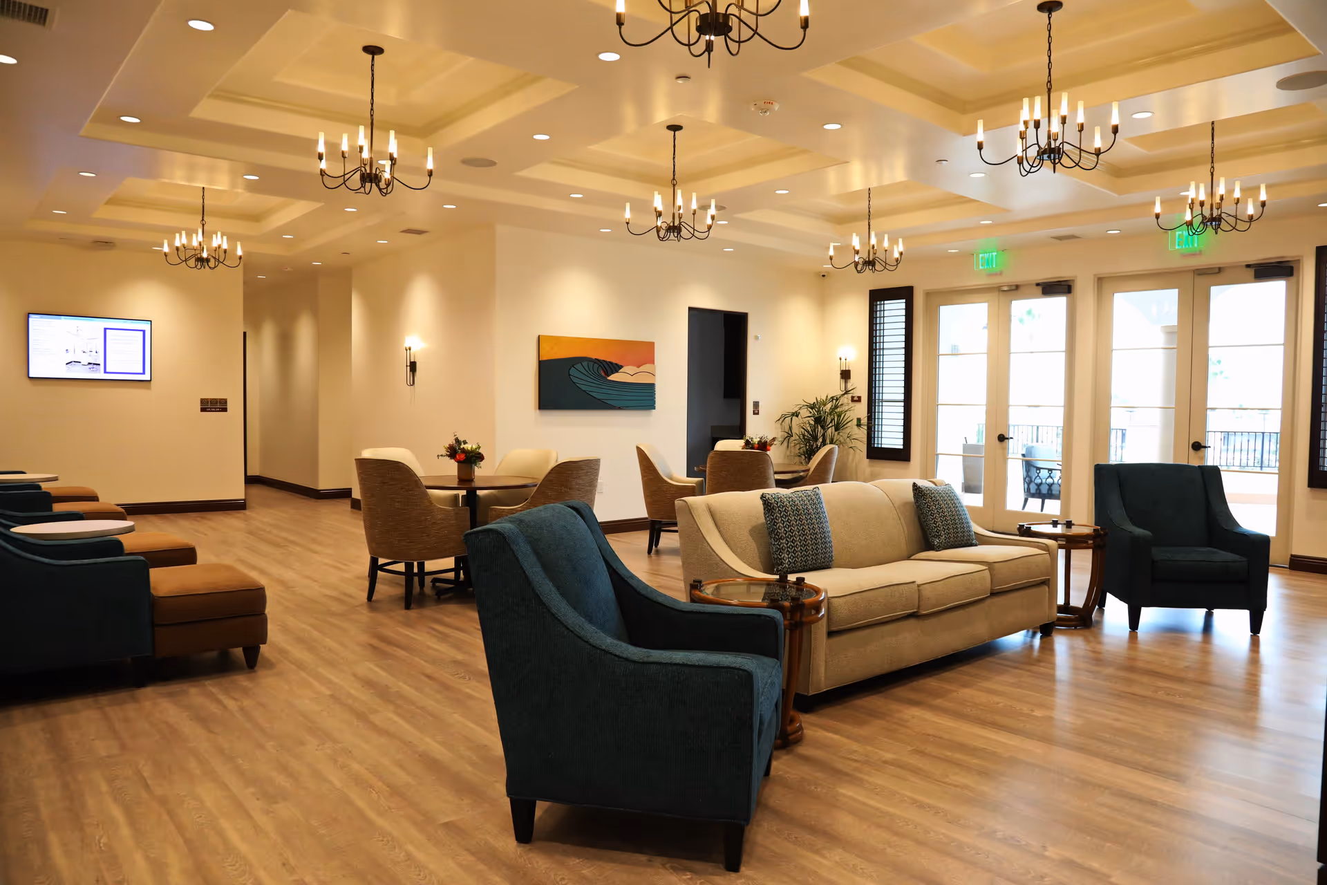 A spacious and well-lit common area with wooden flooring, featuring a beige sofa with patterned cushions, two blue armchairs, round tables with beige chairs, modern chandeliers hanging from a coffered ceiling, wall sconces, a colorful wave-themed painting, and large glass doors leading to an outdoor patio.