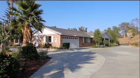 Single-story brick residential building with a garage, surrounded by trees and shrubs under a clear blue sky. A wide concrete driveway curves in front of the building.