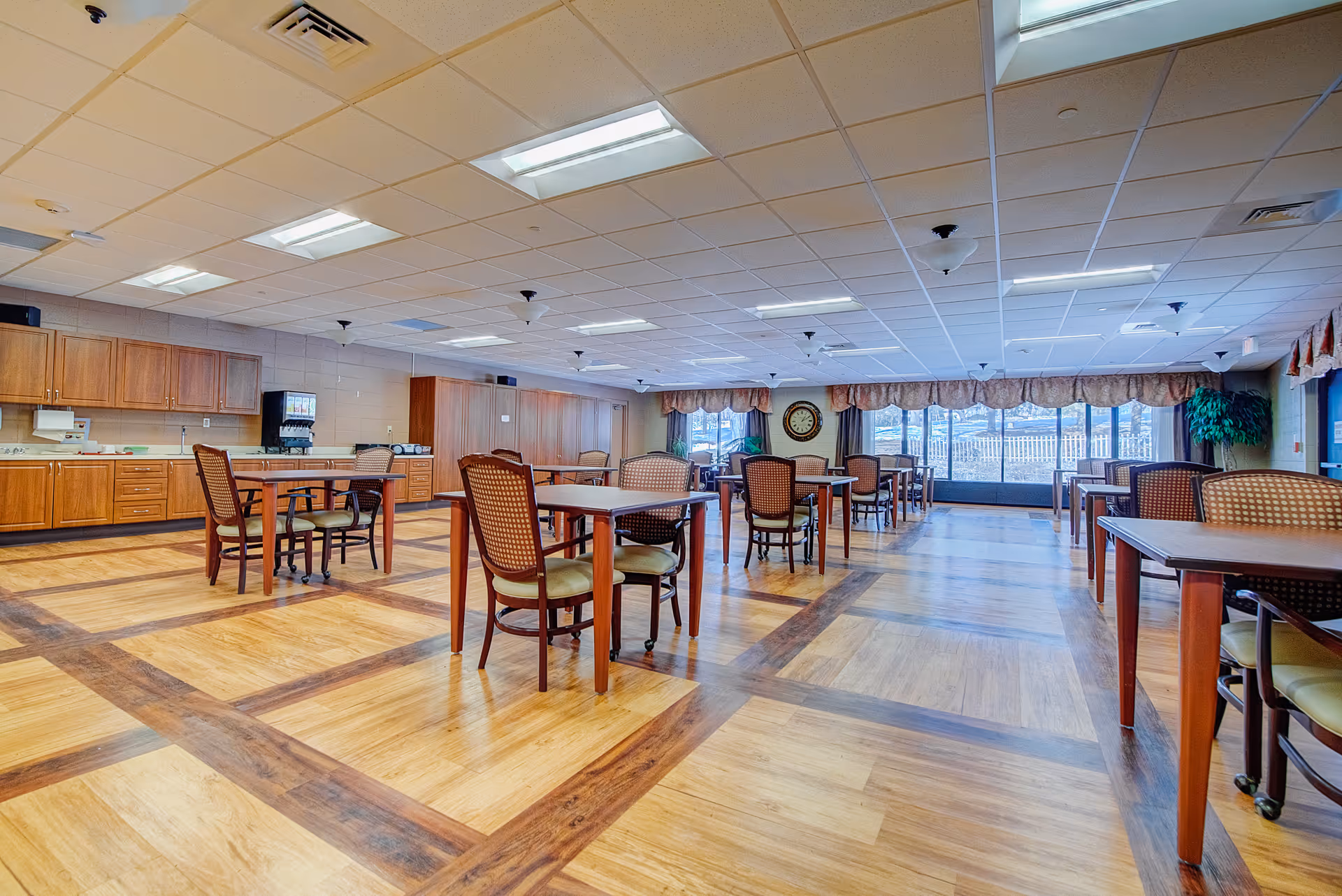 A spacious dining room with multiple wooden tables and chairs arranged neatly on a patterned wooden floor. The room has a drop ceiling with fluorescent lighting and large windows with curtains letting in natural light. There is a clock on the far wall and a beverage dispenser on the left side near wooden cabinets.