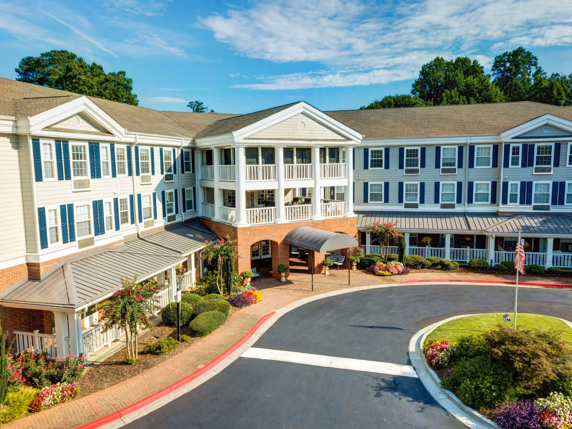 Exterior view of a large, three-story senior living facility building with white siding, blue shutters, and a covered entrance. The building is surrounded by well-maintained landscaping including bushes, flowers, and trees. A circular driveway with a flagpole and a small garden is visible in front of the entrance under a clear blue sky.