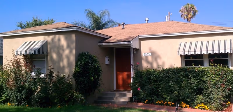 Exterior front view of a single-story beige house with a brown roof, two windows with striped awnings, a red front door, and surrounded by green bushes and flowers under a clear blue sky.