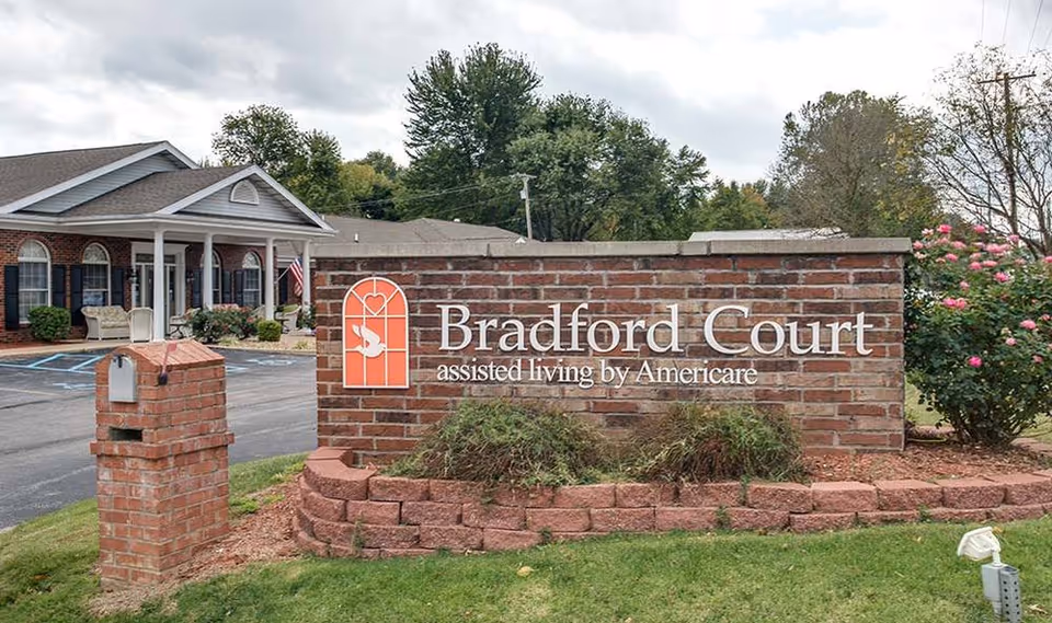 Brick sign for Bradford Court assisted living by Americare in front of a single-story brick building with a covered porch, surrounded by greenery and a parking lot.