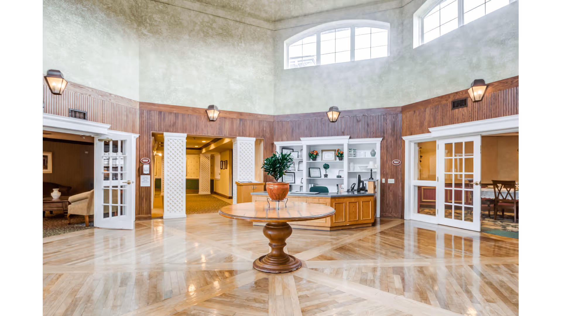 Spacious and well-lit reception area with a round wooden table holding a potted plant in the center. The room features polished wooden floors, wood-paneled walls with sconces, and large windows near the ceiling allowing natural light. There are open double doors on either side leading to other rooms, one with seating and the other with a dining area. A reception desk with shelves and office equipment is positioned against the back wall.