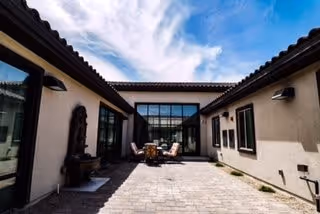 Outdoor courtyard area with stone tile flooring, surrounded by beige walls and windows. There is a seating area with chairs and a table in the center under a clear blue sky with some clouds.