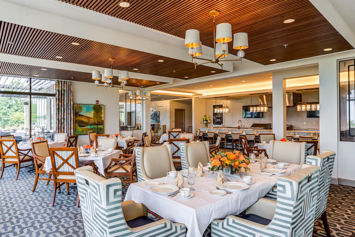 A bright and elegant dining room with multiple tables set with white tablecloths, plates, cups, and napkins. The room features a patterned carpet, wooden chairs with cream cushions, and armchairs with a geometric pattern. There are modern chandeliers hanging from a wooden slatted ceiling, large windows letting in natural light, and a kitchen area with bar seating in the background. A floral centerpiece decorates the table in the foreground.