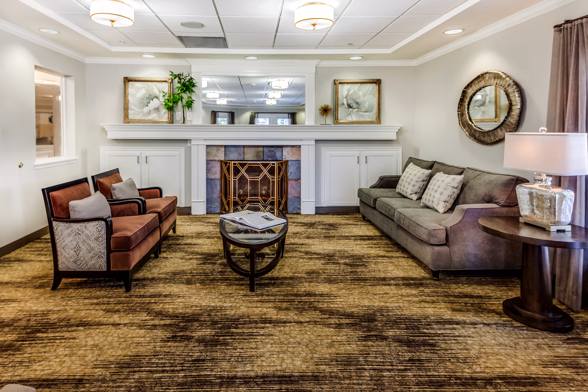 A cozy living room with a brown patterned carpet, a gray sofa with three pillows on the right, two brown armchairs with cushions on the left, a small oval glass coffee table in the center with an open book, a white fireplace with a decorative metal screen, two framed floral paintings above the fireplace, a round mirror on the right wall, and a table lamp on a wooden side table next to the sofa.