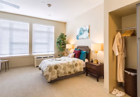 Bright, well-appointed bedroom with a bed, nightstands and lamps, an open closet with robes, and large windows with blinds.