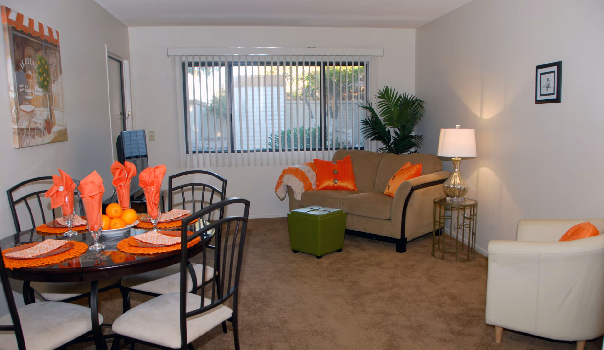 A cozy living and dining area in a retirement community apartment. The dining table is set with orange napkins, placemats, and a bowl of oranges. The living area features a beige loveseat with orange pillows and a throw blanket, a small green ottoman, a white armchair with an orange pillow, a side table with a lamp, a potted plant, and a large window with vertical blinds.