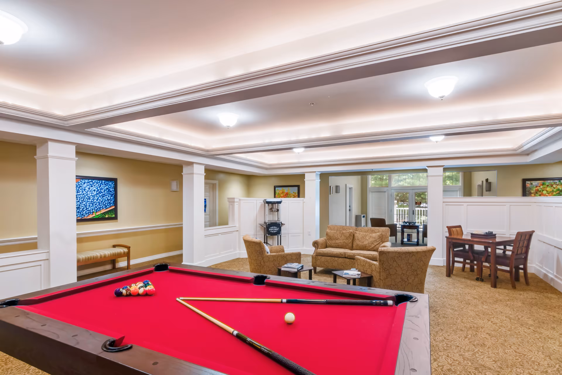 A spacious common room with a red felt pool table in the foreground, two beige patterned sofas with a small coffee table in the middle, a wooden table with four chairs, and bright ceiling lights. The room has white wainscoting, beige walls, and framed artwork on the walls. Large windows and glass doors in the background let in natural light.