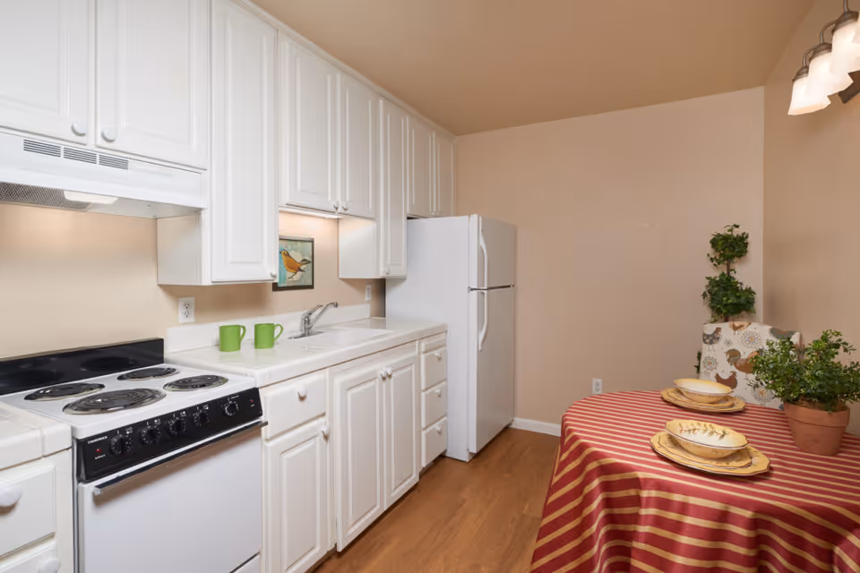 A clean kitchen with white cabinets, a white refrigerator, an electric stove with four burners, and a white countertop with a sink. Two green mugs are placed on the counter. To the right, there is a small dining area with a round table covered with a red and beige striped tablecloth, set with plates and bowls. There are two potted plants and a chair with a patterned cover featuring roosters.