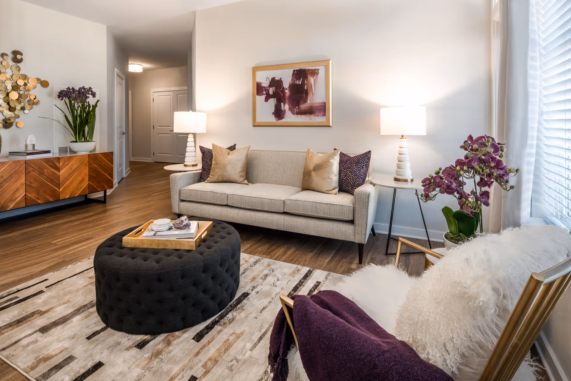 A modern living room with a beige sofa adorned with gold and purple pillows, two white table lamps on either side, a black tufted ottoman with a gold tray on top, a wooden sideboard with a plant and decorative items, a white furry chair with a purple throw, and a large window with white blinds letting in natural light.
