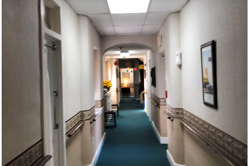 A long indoor hallway with green carpet and beige walls featuring a decorative border. Handrails run along both sides of the hallway. There are doors and framed pictures on the walls, with chairs and a small table with a flower arrangement visible further down the hall. The ceiling has white tiles with fluorescent lighting.