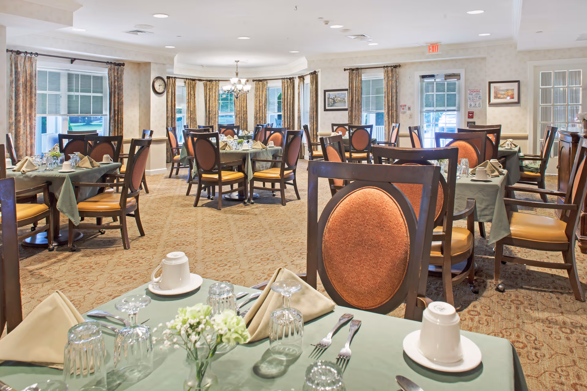 Spacious dining room set for a meal with multiple tables, upholstered chairs, place settings, and windows.