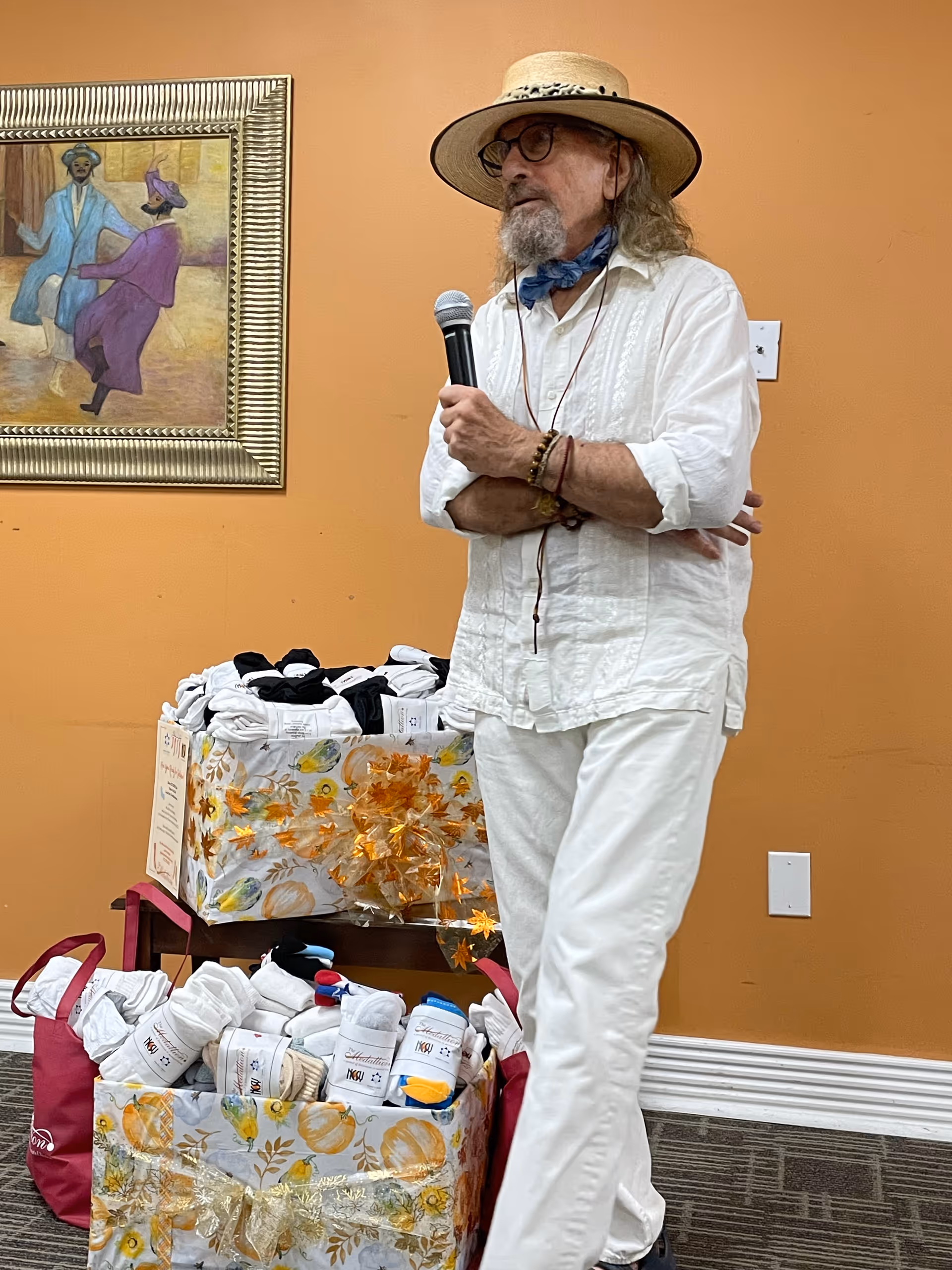An elderly man wearing a white outfit, a straw hat, glasses, and a blue neck scarf is holding a microphone and standing in front of an orange wall. Behind him are two large gift boxes decorated with autumn-themed wrapping paper and filled with rolled white socks. A framed painting of two people dancing hangs on the wall.
