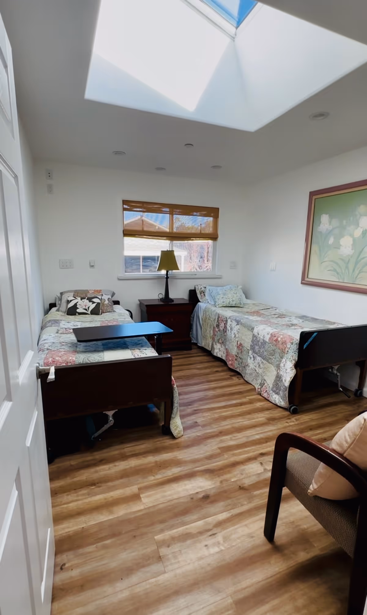 A bright bedroom with two single beds covered in patchwork quilts, a wooden nightstand with a lamp between the beds, a window with a brown blind, a skylight above, and a framed floral painting on the wall. There is also a cushioned chair with a pillow in the corner.