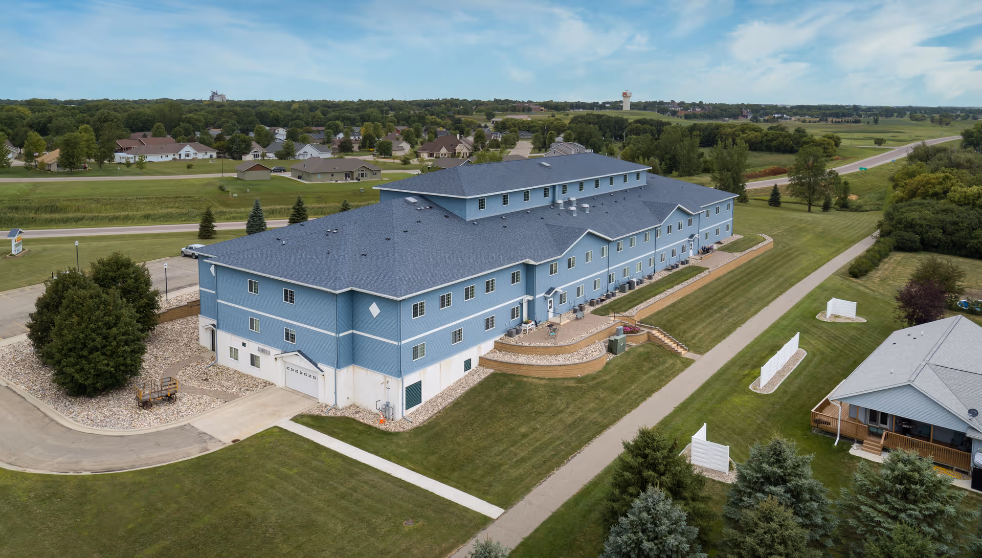 Aerial view of a large blue multi-story senior living building with lawns, walkways, and nearby houses.